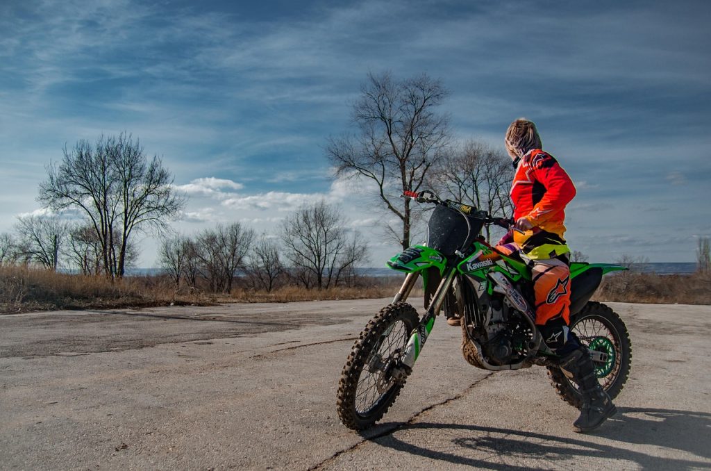 A rider in bright red and orange gear sits on a green dirt bike, looking off into the distance on an open concrete area with bare trees under a blue sky.