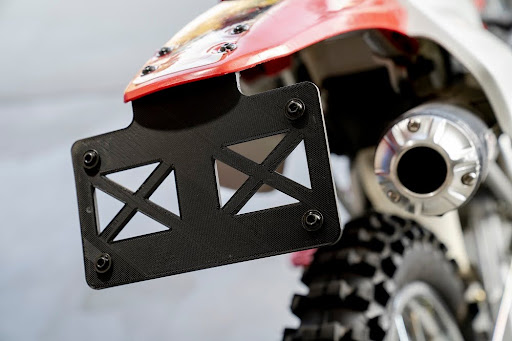 This image shows the rear end of a dirt bike featuring a knobby tire, an aftermarket exhaust, and a custom lightweight license plate holder with cutout patterns for reduced weight and airflow.