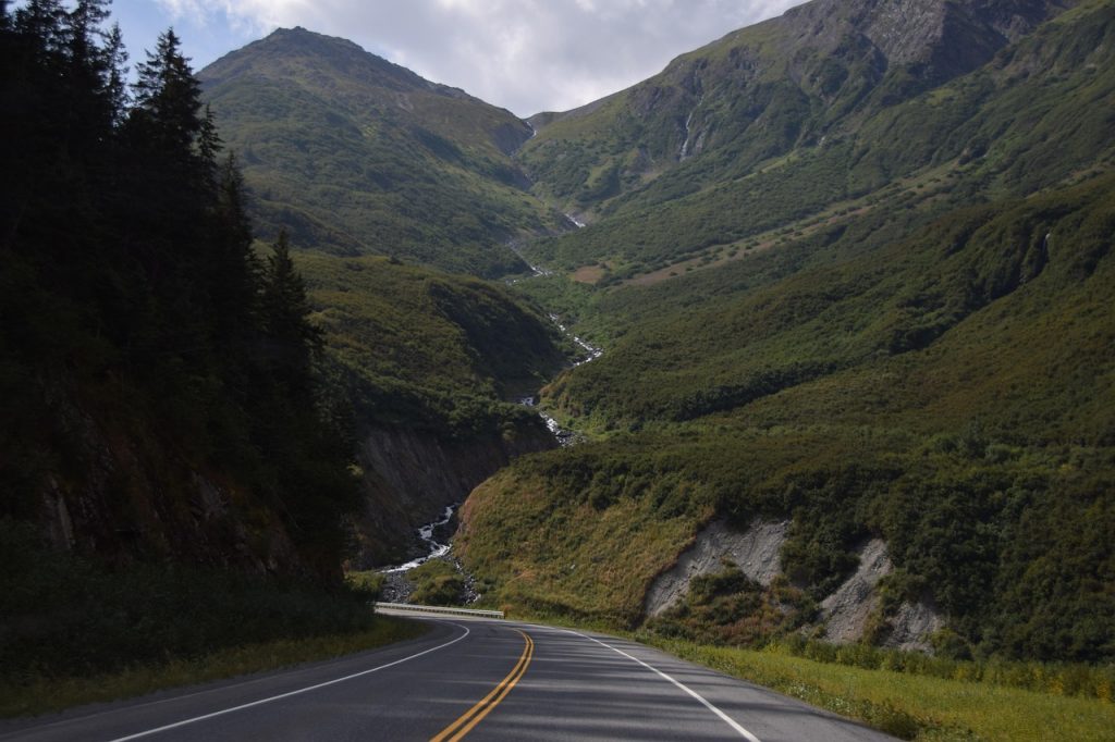 A winding paved road curves through a lush green mountain valley, with forested slopes, rocky ridges, and a cascading stream running down the center.