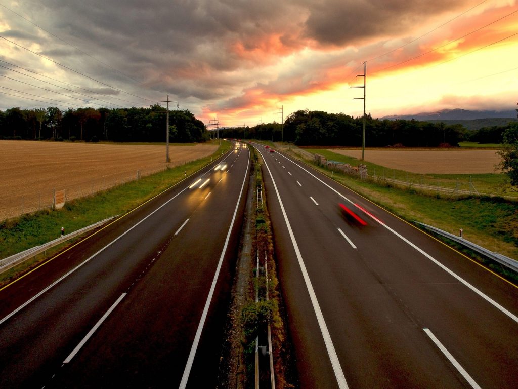A divided highway glows under a dramatic sunset as cars with plates speed freely toward their destinations, embodying the freedom of the open road.