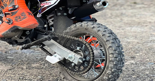 This image shows the rear wheel of a small dirt bike with a knobby off-road tire, visible chain drive, and an orange motocross boot on the footpeg.