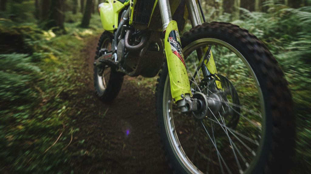 Close-up of a neon green dirt bike's front wheel and suspension on a muddy, mossy forest trail.