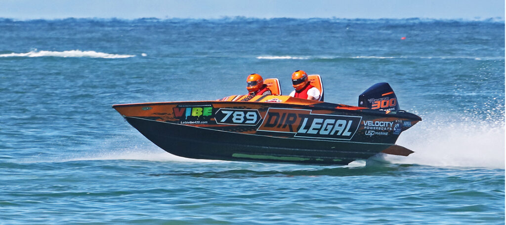 Orange and black 'Dirt Legal' powerboat, number 789, with two people in orange helmets, racing across blue ocean waves, kicking up spray. 'Velocity Powerboats' and 'Mercury Racing 300' logos are visible on the boat and engine. The boat is slightly airborne due to speed.
