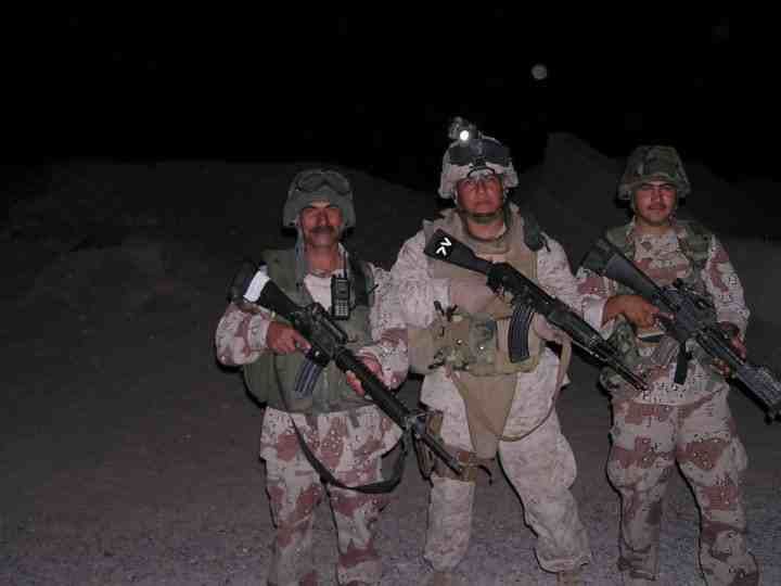 Three U.S. soldiers in full combat gear stand together at night in a desert environment, armed and ready, with serious expressions under a dark sky