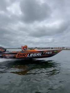 High-speed powerboat 'Dirt Legal 789' with a 'Velocity Powerboats' logo, skimming low over dark water under a dramatic grey sky. The driver, wearing a bright orange helmet, is visible inside the cockpit.