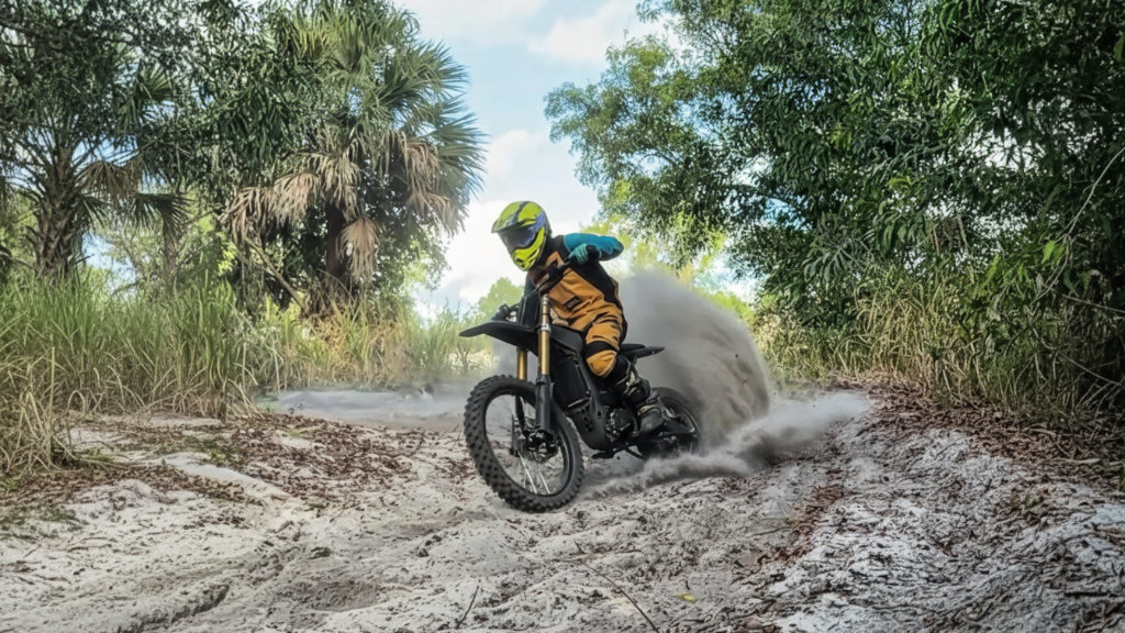 E-Moto rider on a performance-upgraded Sur Ron electric dirt bike performing an aggressive off-road wheelie and kicking up dirt, showcasing GritShift parts.