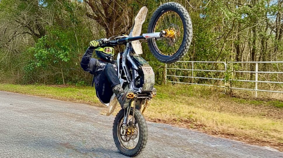 E-Moto rider in full gear performing a high wheelie on a modified electric dirt bike on a paved road, demonstrating GritShift performance.