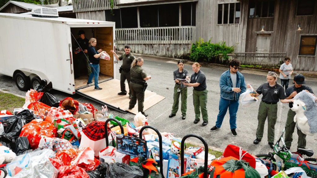 Volunteers and law enforcement officers unloading bags of toys from a white trailer for a charity event, with large piles of donated gifts in the foreground
