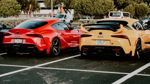 Rear view of a red and a yellow Toyota GR Supra MK5 parked in a lot, showcasing custom spoilers and Florida license plates.