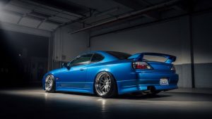 Modified blue Nissan Silvia S15 JDM sports car with stanced suspension and deep dish chrome wheels parked in a dark garage with dramatic lighting.