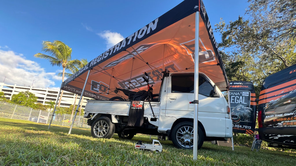 A white Japanese Kei truck parked under a branded Dirt Legal pop-up tent for street legal vehicle registration at an outdoor event.