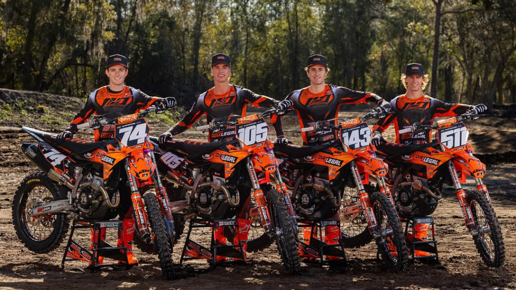 Four professional motocross riders in matching orange and black racing jerseys stand behind their Dirt Legal sponsored KTM motorcycles in a wooded outdoor setting.